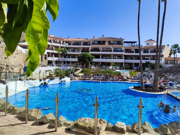 Sunny hotel pool with lounging guests and greenery.